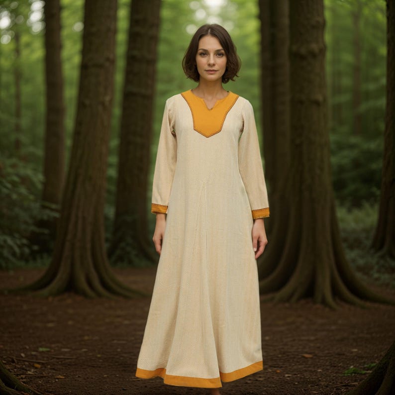 A woman stands in a forest, wearing a long, cream-colored dress with a yellow V-neck collar and yellow trim on the cuffs and hem.