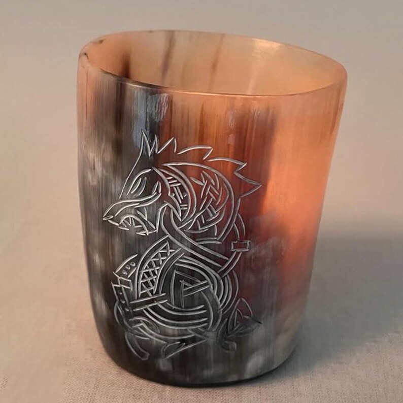 May include: A horn drinking vessel with a carved wolf design. The cup features a colour gradient from dark grey to light brown and orange. The wolf is depicted in a detailed Celtic knot style.