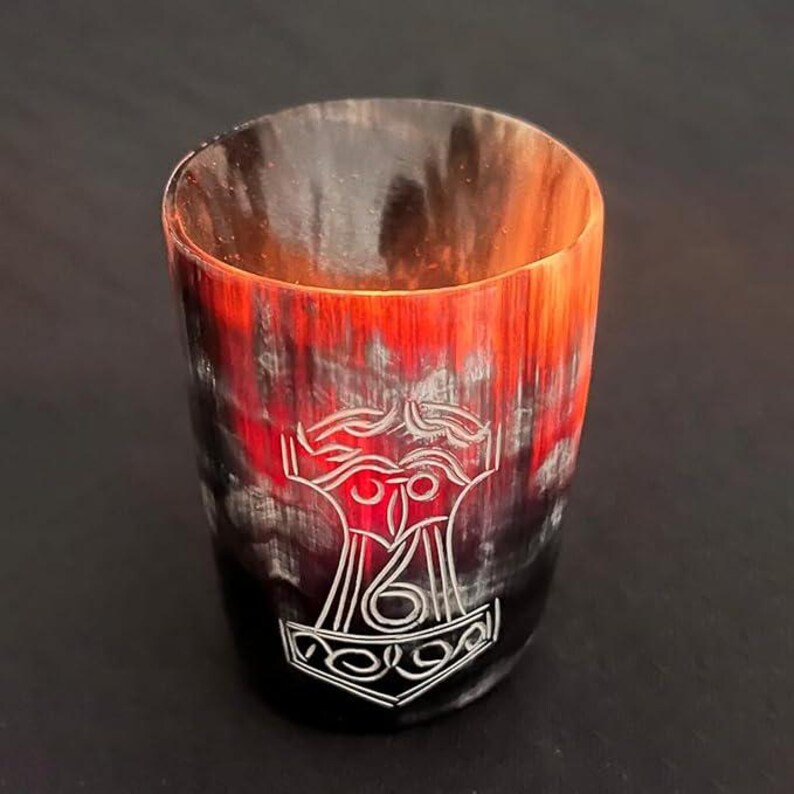 May include: A cylindrical drinking glass with a natural, textured appearance, displaying a colour gradient from black to red-orange. The front of the glass features a white, etched image of Thor's hammer, Mjolnir. The glass has a smooth, polished finish.