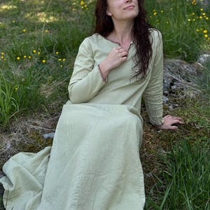 May include: A light green long-sleeved dress with a V-neckline and black stitching detail. The dress is made of a soft, flowing fabric and is suitable for historical reenactments or casual wear.