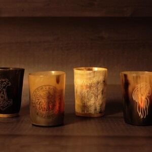 May include: Four Viking-style drinking horns with engraved designs. The horns range in colour from dark brown to light tan. Each horn features a unique etched image, including a hammer, a sun-like emblem, a warrior, and a long-haired figure.