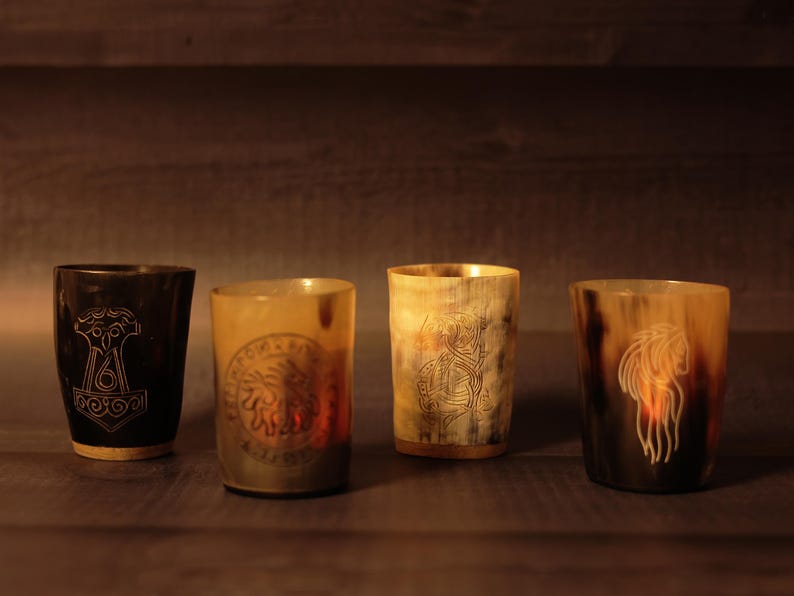 May include: Four Viking-style drinking horns with engraved designs. The horns range in colour from dark brown to light tan. Each horn features a unique etched image, including a hammer, a sun-like emblem, a warrior, and a long-haired figure.