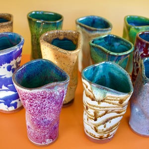 Peut inclure: Collection de gobelets en céramique faits à la main de différentes couleurs, dont le bleu, le vert, le violet et le marron. Chaque gobelet a un glaçage et une forme uniques, avec un intérieur turquoise. Les gobelets sont disposés sur une surface orange.