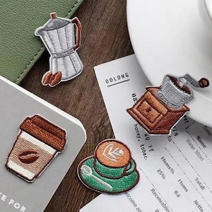 May include: Embroidered coffee-themed patches. Designs include a coffee maker, coffee grinder, takeaway cup, and a coffee cup with latte art. Patches are in brown, green, and silver, placed on a notebook and a receipt.
