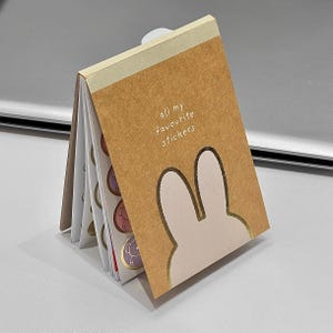 May include: A small, open notebook with a brown cover featuring a bunny silhouette and the text "all my favourite stickers." The notebook contains several pages of colorful stickers with various designs, including animals. The notebook is open, displaying the stickers.