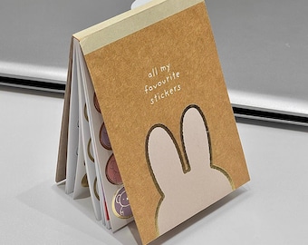 May include: A small, open notebook with a brown cover featuring a bunny silhouette and the text "all my favourite stickers." The notebook contains several pages of colorful stickers with various designs, including animals. The notebook is open, displaying the stickers.