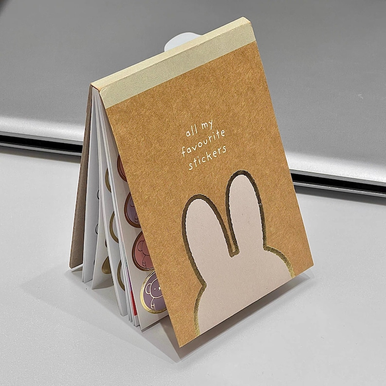 May include: A small, open notebook with a brown cover featuring a bunny silhouette and the text "all my favourite stickers." The notebook contains several pages of colorful stickers with various designs, including animals. The notebook is open, displaying the stickers.