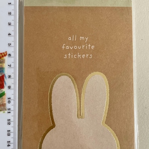 May include: A brown sticker book with the text "all my favourite stickers" in white. The cover features a bunny ear design with gold trim. The book is sealed in clear plastic.