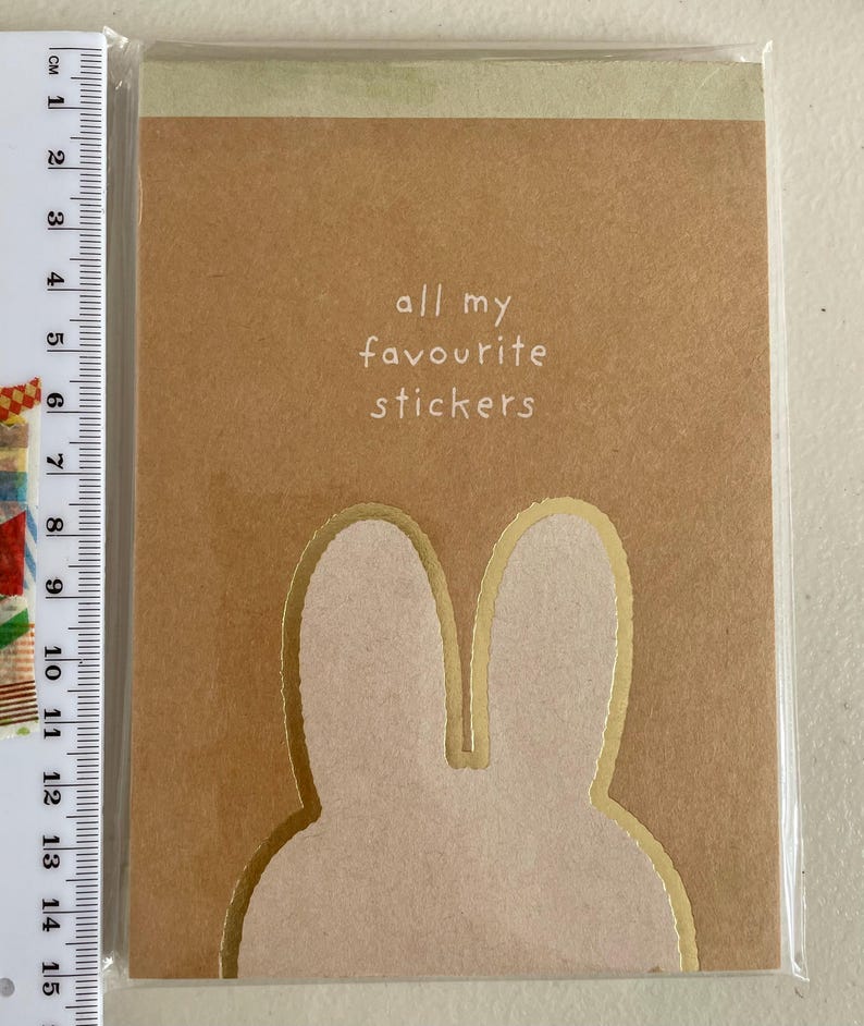 May include: A brown sticker book with the text "all my favourite stickers" in white. The cover features a bunny ear design with gold trim. The book is sealed in clear plastic.