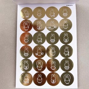 May include: Sheet of gold foil stickers featuring white line art illustrations of a bunny character. Each sticker is a circle with a different pose of the bunny. The stickers are on a white background.