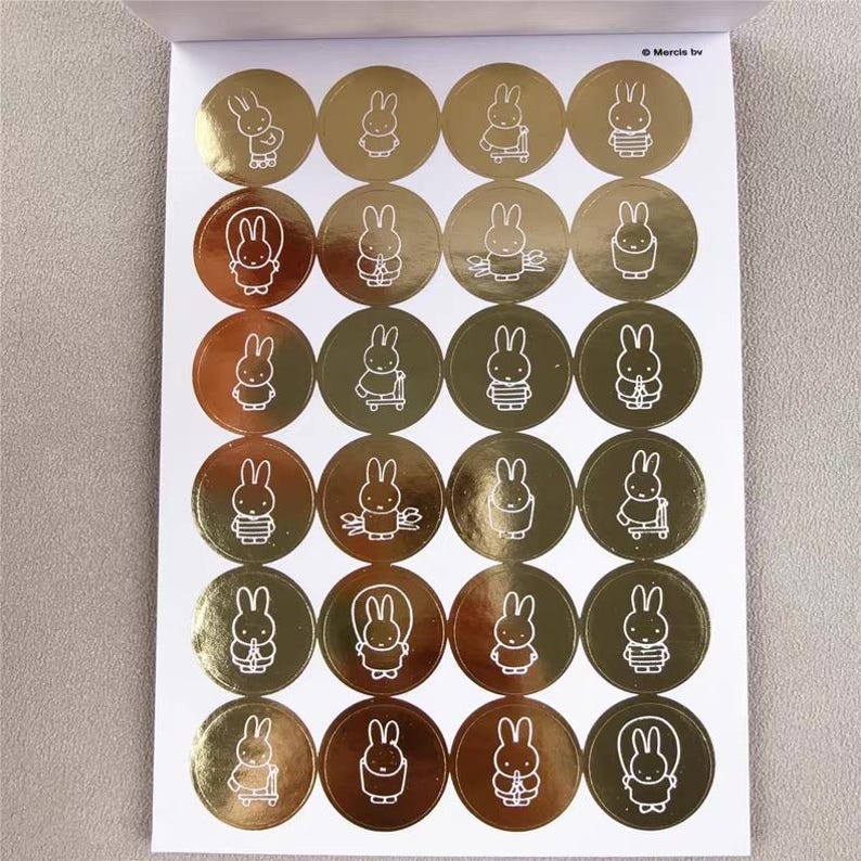 May include: Sheet of gold foil stickers featuring white line art illustrations of a bunny character. Each sticker is a circle with a different pose of the bunny. The stickers are on a white background.