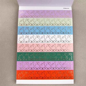 May include: A sheet of stickers featuring rows of cartoon bunnies in various colors. Each bunny has a gold outline. The background colors include purple, green, blue, white, pink, and orange. The stickers are on a white background.