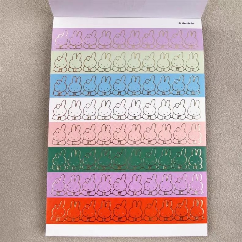 May include: A sheet of stickers featuring rows of cartoon bunnies in various colors. Each bunny has a gold outline. The background colors include purple, green, blue, white, pink, and orange. The stickers are on a white background.