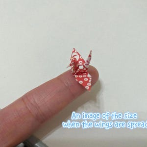 Könnte beinhalten: Ein winziger Origami-Kranich, rot mit weißem Blumenmuster, sitzt auf einer Fingerspitze. Die Flügel des Kranichs sind ausgebreitet und zeigen seine filigrane Papierkonstruktion. Text auf dem Bild: "An image of the size when the wings are spread."