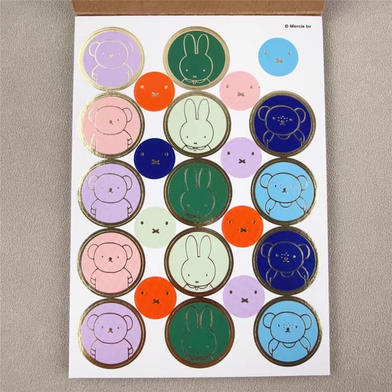 May include: A sheet of colorful stickers featuring cartoon bears, bunnies, and other circular designs. The stickers are in various colors, including pink, purple, green, blue, and orange, with gold foil accents. The stickers are on a white background.