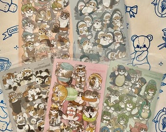 Puede incluir: Cinco hojas de pegatinas con varios personajes de animales de dibujos animados con diferentes atuendos. Las pegatinas están en un embalaje de plástico transparente. Los personajes incluyen gatos, perros y pandas. El fondo es de tela estampada azul claro y blanco.