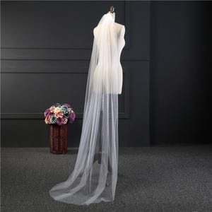 May include: A long, white tulle wedding veil with a single layer. The veil is draped over a white dress form.