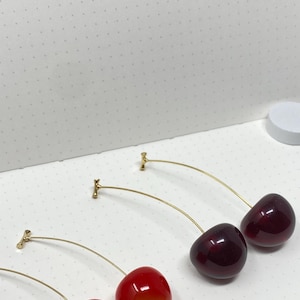 Puede incluir: Un par de pendientes de cereza de oro con cerezas rojas y rojo oscuro. Las cerezas son de vidrio y tienen un acabado brillante.