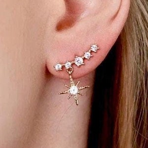 May include: Gold-toned ear climber earring with a series of small, clear gemstones ascending the ear. A dangling starburst charm with clear gemstones hangs below. The earring is worn on a person's ear.