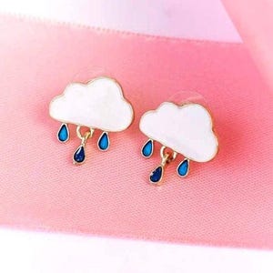 Puede incluir: Pendientes de botón con forma de nube. Cada pendiente presenta una nube blanca con ribete dorado y tres gotas azules colgantes. Sobre un fondo rosa con una cinta rosa.