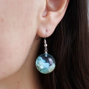 Może przedstawiać: Okrągłe kolczyki z żywicy w kolorze niebieskim i białym z haczykiem ze srebra. Kolczyki mają wzór chmurnego nieba.