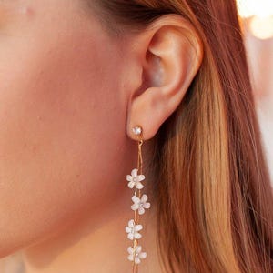 Puede incluir: Pendientes de aro dorados con una cadena delicada y pequeños detalles de flores blancas.