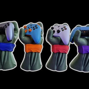 May include: Four green hand sculptures, each with a different colored wristband: blue, orange, red, and purple. Each hand holds a different colored video game controller: black, blue, lavender, and blue. The background is black.