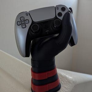 May include: A black and gray video game controller is held by a black hand-shaped stand with red and black accents. The controller has a silver faceplate and black buttons. The stand has a wrist-like design with red bands.