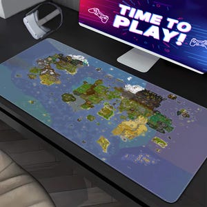 Op de afbeelding: Een grote rechthoekige gaming muismat met een gedetailleerd kaartontwerp met landmassa's en water. De mat ligt op een bureau met een computermonitor met "TIME TO PLAY!" en een VR-headset.
