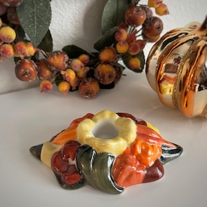 May include: A ceramic candle holder in the shape of a flower with a yellow center and colorful petals in shades of orange, red, and green. A decorative fall wreath and a gold pumpkin ornament are in the background.