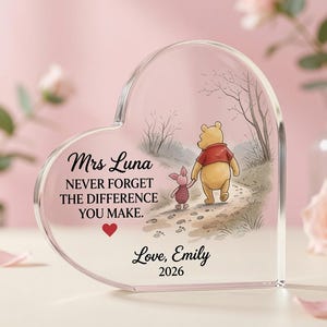 May include: A heart-shaped clear acrylic plaque featuring a printed illustration of Winnie the Pooh and Piglet walking on a path. The text reads: Mrs Luna, NEVER FORGET THE DIFFERENCE YOU MAKE., Love, Emily, 2026, with a small red heart icon.