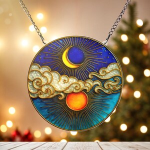 Puede incluir: Colgante redondo de cristal manchado con el sol y la luna, con un fondo azul, detalles dorados y una cadena plateada. El diseño presenta una luna creciente, el sol y nubes. El colgante mide aproximadamente 10 cm de diámetro.