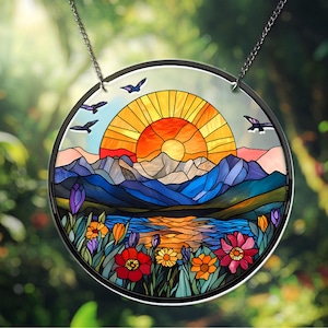 May include: Round stained glass window art depicting a sunset over mountains and a lake, with colorful flowers in the foreground. The sun is a vibrant orange and yellow, and the mountains are shades of blue and gray. Three birds are flying in the sky.