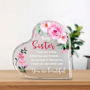 Può includere: Un blocco di acrilico trasparente a forma di cuore con le parole "To my Sister" in corsivo rosso, circondato da accenti floreali rosa. Il blocco include il testo "I love you today, tomorrow and forever, As you look in the mirror, I hope you see what I see, You are beautiful."