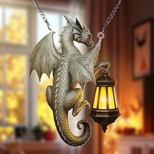 Könnte beinhalten: Eine silberfarbene Drachenskulptur mit ausgebreiteten Flügeln, die eine Laterne mit warmem gelbem Schein hält. Der Drache ist an einer Kette aufgehängt und zeigt komplizierte Details und ein Fantasy-Thema.