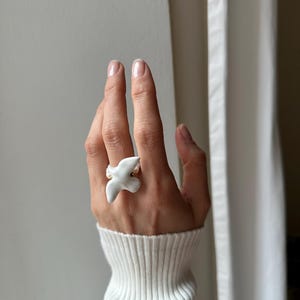 May include: A white ceramic ring in the shape of a bird, with gold-colored accents. The ring is worn on a hand, with a white ribbed sweater in the background. The bird ring is a unique piece of jewelry.