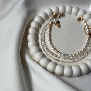 May include: A collection of delicate necklaces with pearls and gold accents, displayed on a white, beaded tray. The necklaces vary in style, featuring small pearls, clear crystals, and gold chains. The tray sits on a white, textured surface.