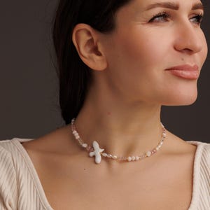 May include: A delicate necklace featuring a white ceramic bird pendant. The necklace is adorned with small pearls, pink beads, and clear crystals, creating a subtle, elegant design. The necklace is a beautiful piece of jewelry.