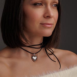 May include: A silver heart pendant necklace with a black choker style cord. The necklace is worn by a person with dark hair and a white fluffy sweater.