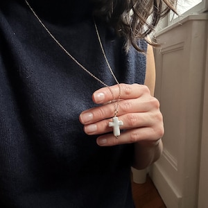 Op de afbeelding: Een goudkleurige ketting met een witte kruisvormige hanger. Het kruis wordt vastgehouden door een persoon die een marineblauw mouwloos topje draagt. Een eenvoudig, elegant accessoire.