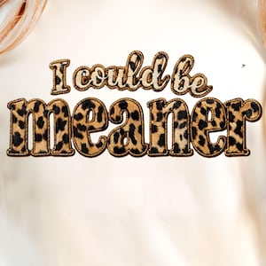 Peut inclure: Sweat-shirt crème avec l'inscription "I could be meaner" dans un motif léopard. Le texte est centré sur le devant du t-shirt. Les manches sont retroussées. Le modèle porte un jean bleu.