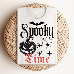 Peut inclure: T-shirt blanc avec un motif sur le thème d'Halloween. Le motif comprend les mots "Spooky Time" en noir et rouge, une citrouille d'Halloween, une chauve-souris, une araignée et des étoiles. Le t-shirt est plié sur une surface circulaire tissée.
