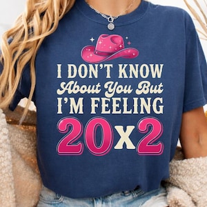 Peut inclure: T-shirt bleu marine avec l'inscription "I DON'T KNOW ABOUT YOU BUT I'M FEELING 20X2" en blanc et rose. Le graphisme comprend un chapeau de cow-boy rose. Le t-shirt est porté par une personne.