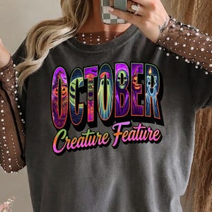 Peut inclure: Un t-shirt gris anthracite à manches longues avec des manches en maille transparente ornées de petites perles blanches. L'avant présente le texte "OCTOBER" avec des illustrations colorées d'Halloween dans chaque lettre et "Creature Feature" en dessous.
