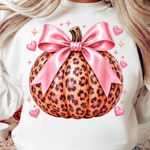 Peut inclure: Un sweat-shirt blanc à manches longues avec un motif central représentant une citrouille orange à imprimé léopard noir. La citrouille est ornée d'un grand nœud en satin rose brillant sur le dessus, entouré de petits cœurs et d'étoiles roses.