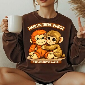 May include: A brown sweatshirt featuring a graphic of two hugging cartoon monkeys. The text "HANG IN THERE, PUNCH" is above the monkeys, and "YOU ARE NEVER ALONE" is below. The sweatshirt is a solid brown color.