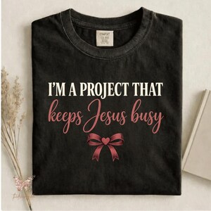 May include: Black t-shirt with the text "I'M A PROJECT THAT keeps Jesus busy" in white and pink script. A pink bow graphic is below the text. The shirt has a crew neck and a Comfort Colors tag.
