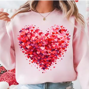 Peut inclure: Un sweat-shirt rose clair orné d'un grand cœur composé de nombreux cœurs rouges, roses et violets. Le cœur est placé au centre du devant du sweat-shirt, sur le thème de la Saint-Valentin.