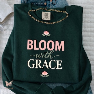Peut inclure: Sweat-shirt vert foncé avec l'inscription "BLOOM with GRACE" en rose et blanc, ornée de motifs floraux. Un collier chaîne doré est posé sur le sweat-shirt. Le sweat-shirt est présenté sur une surface blanche avec un jean bleu.