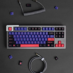 Könnte beinhalten: Eine silberne mechanische Tastatur mit blauen und roten Tastenkappen. Die Tastatur hat japanische Schriftzeichen und das Wort "Ninja" aufgedruckt. Ein gewendeltes graues Kabel und zusätzliche Tastenkappen sind ebenfalls sichtbar.